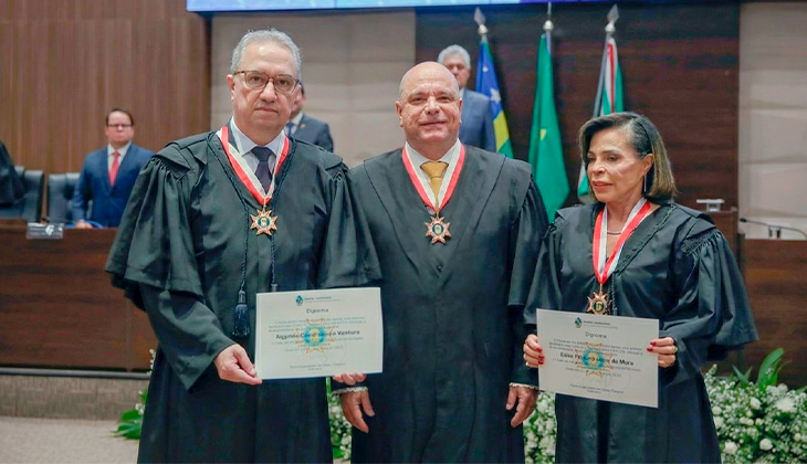 A foto mostra três pessoas, duas delas segurando certificados, todos vestindo togas pretos e usando no pescoço uma medalha em formato de estrela, presa por uma faixa vermelha e branca.  Eles estão em um ambiente formal, que parece ser um auditório institucional, com uma mesa de autoridades ao fundo e bandeiras do Brasil e de estados brasileiros atrás deles.  À esquerda, o desembargador Augusto Ventura. Homem de óculos, cabelos grisalhos curtos e expressão séria. Ele segura um certificado com as duas mãos.  Ao centro, o presidente do TJGO, Leandro Crispim. Homem careca, sorrindo levemente, também de toga e medalha, mas sem segurar nenhum documento.  À direita, a desembargadora Edna Horta. Mulher de cabelos curtos escuros, segurando um certificado semelhante ao do homem da esquerda.  Todos estão bem posicionados para uma foto oficial, sugerindo que participaram de uma cerimônia de posse, homenagem ou entrega de honrarias.