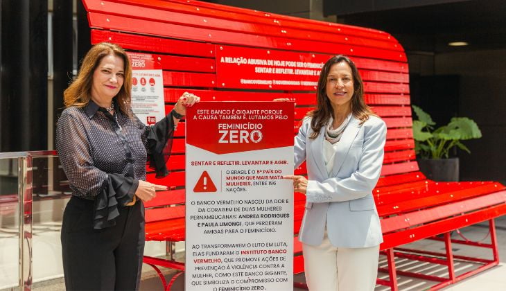 Duas mulheres adultas estão de pé, uma ao lado da outra, segurando um grande painel informativo. Uma é a desembargadora Alice Teles de Oliveira e servidora do TJGO, Luciene Camargo, esposa do presidente do Tribunal. Elas posam sorrindo em frente a um banco público de cor vermelha intensa, que é largo e chamativo. O banco traz inscrições sobre o combate ao feminicídio.  A mulher à esquerda tem cabelos castanhos ondulados, veste uma blusa preta transparente com mangas longas e calça preta. A mulher à direita tem cabelos castanhos lisos, veste um conjunto claro composto por blazer e calça, com uma blusa branca por baixo.  Entre elas, está o painel branco e vermelho que fala sobre a iniciativa “Feminicídio Zero”. Nele, há textos explicando a importância de refletir e agir contra a violência contra mulheres, citando estatísticas e a origem do projeto do “Banco Vermelho”.  O cenário é interno, provavelmente um saguão de edifício, com iluminação clara e algumas plantas decorativas ao fundo.