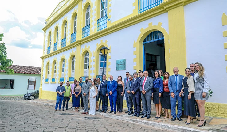 Imagem em formato horizontal, ao ar livre, durante o dia. Em frente a um prédio histórico de dois andares, um grupo numeroso de pessoas posa para foto. O edifício tem fachada predominantemente branca com detalhes em amarelo, janelas em arco e sacadas com grades azuis, além de uma porta central também em arco. O prédio aparenta ser antigo e restaurado, com estilo arquitetônico clássico.  À frente da fachada, cerca de vinte pessoas estão alinhadas lado a lado, formando uma fila. Elas vestem trajes formais e sociais: homens usam, em sua maioria, ternos em tons escuros com camisas claras e gravatas; mulheres usam vestidos, saias, calças sociais e blazers em cores variadas, como preto, branco, azul e bege. A maioria está sorrindo e olhando em direção à câmera, com postura ereta.  O chão é de pedras irregulares, típico de calçamento histórico. À esquerda da imagem, é possível ver parte de uma árvore, um carro estacionado e uma construção simples ao fundo. O céu aparece parcialmente nublado, contribuindo para uma iluminação natural suave na cena.