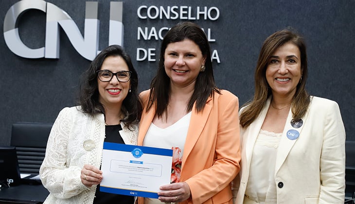 Foto colorida, em ambiente interno institucional. Três mulheres adultas estão em pé, lado a lado, sorrindo para a câmera. Ao fundo, há uma parede escura com o logotipo e a inscrição “CNJ – Conselho Nacional de Justiça”, indicando que a cena ocorre em uma sala oficial do órgão.  À esquerda, uma mulher de pele clara, cabelos escuros, lisos e na altura dos ombros. Ela usa óculos de grau com armação escura, batom vermelho, blusa clara e um blazer branco rendado. Ela segura, com as duas mãos, um certificado ou documento oficial de cor branca e azul.  No centro, uma mulher de pele clara, cabelos castanhos escuros, lisos e longos, soltos. Ela veste um blazer de tom pêssego ou salmão sobre uma blusa clara estampada. Está posicionada levemente à frente das outras duas, também sorrindo.  À direita, uma mulher de pele clara, cabelos castanhos claros, lisos e longos, soltos. Ela usa um blazer branco sobre uma blusa clara e tem um broche redondo preso à lapela do blazer. Também sorri para a câmera.  A iluminação é uniforme e profissional, e a imagem transmite um clima formal e institucional, associado a reconhecimento ou entrega de certificação.