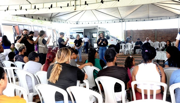 Imagem colorida, em ambiente aberto e coberto por uma grande tenda branca. No centro e em primeiro plano, há várias pessoas sentadas em cadeiras plásticas brancas, dispostas em fileiras, vistas principalmente de costas e de perfil. O público é diverso, com adultos, idosos e algumas crianças, usando roupas casuais.  Ao fundo, à esquerda, algumas pessoas estão em pé, algumas segurando câmeras fotográficas ou celulares, aparentemente registrando o evento. No centro do fundo, há um veículo branco tipo van, com identificação de serviço público de saúde, estacionado sob a tenda. Próximo à van, duas mulheres em pé falam ao público; uma delas segura um microfone, indicando que está conduzindo ou participando de uma apresentação ou orientação.  À direita do fundo, há mais cadeiras brancas, algumas vazias, e uma mesa coberta com toalha clara. O ambiente sugere uma ação comunitária ou evento público, possivelmente de saúde ou informação, realizado durante o dia, com iluminação natural.