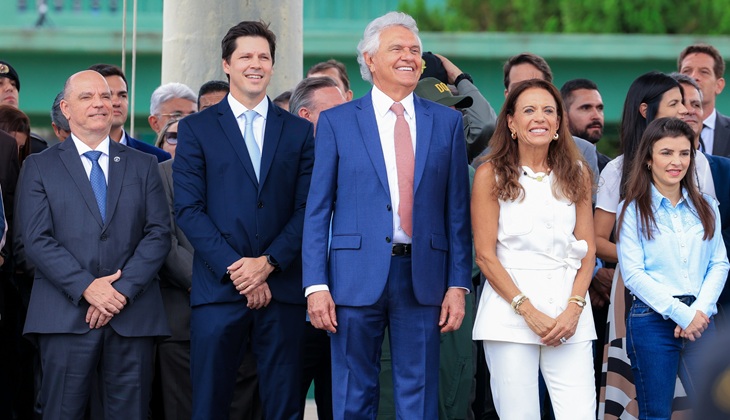 Fotografia colorida, em ambiente externo, durante um evento oficial. No centro da imagem há um grupo de pessoas adultas, em pé, alinhadas lado a lado, aparentando assistir a uma cerimônia. A maioria veste roupas formais. Destaca-se um homem de cabelo grisalho, usando terno azul e gravata clara, sorrindo e olhando para frente. À sua esquerda e à direita estão outros homens de terno escuro e mulheres com trajes sociais claros e escuros. Uma mulher à direita usa roupa branca sem mangas; outra, mais à direita, veste camisa azul-clara e calça jeans. Ao fundo, vê-se mais pessoas, algumas parcialmente desfocadas, e uma estrutura verde, possivelmente de um prédio público. A expressão geral do grupo é de atenção e cordialidade, sugerindo um momento solene ou comemorativo.