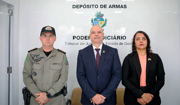 Fotografia em ambiente interno, bem iluminado, com fundo branco. Ao centro do fundo há a inscrição “DEPÓSITO DE ARMAS” e, abaixo, “PODER JUDICIÁRIO – Tribunal de Justiça do Estado de Goiás”, acompanhada de um brasão oficial.  Em primeiro plano, três pessoas estão de pé, lado a lado, voltadas para a câmera, com postura formal e mãos à frente do corpo:  À esquerda, uma pessoa usando uniforme da Polícia Militar, em tom bege, com insígnias nos ombros, distintivos no peito e um boné preto com emblema. Usa cinto tático com equipamentos e mantém expressão neutra.  Ao centro, uma pessoa vestindo terno azul-escuro, camisa clara e gravata vinho, com um pequeno broche na lapela. Está com expressão serena e postura formal.  À direita, uma pessoa vestindo blazer preto sobre blusa em tom salmão, também com broche na lapela, cabelo escuro solto e expressão séria.