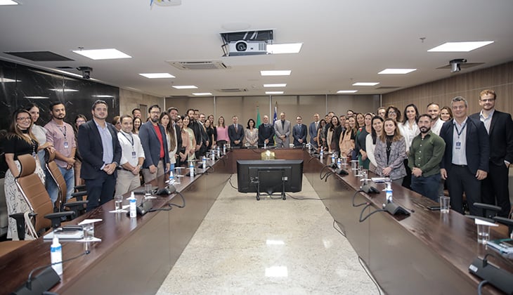 Imagem em ambiente interno, em uma sala de reuniões ampla e bem iluminada. No centro, há uma grande mesa retangular de madeira escura, com microfones, copos de água, garrafas e fios distribuídos ao longo de sua extensão. Na extremidade da mesa, vê-se um monitor de televisão voltado para o interior da sala.  Ao redor da mesa, em pé, estão aproximadamente cinquenta pessoas adultas, de diferentes gêneros, idades aparentes e tons de pele. A maioria veste roupas formais ou sociais, como ternos, blazers, camisas e vestidos, alguns com crachás de identificação. As pessoas estão dispostas de ambos os lados da mesa e também ao fundo da sala, olhando para a câmera, muitas com expressão neutra ou sorrindo levemente.  O ambiente possui paredes em tons neutros, uma delas com acabamento em pedra escura. No teto, há iluminação embutida e um projetor central. O piso é claro e polido, refletindo a luz do ambiente. A imagem transmite a ideia de um encontro institucional, reunião oficial ou evento corporativo.