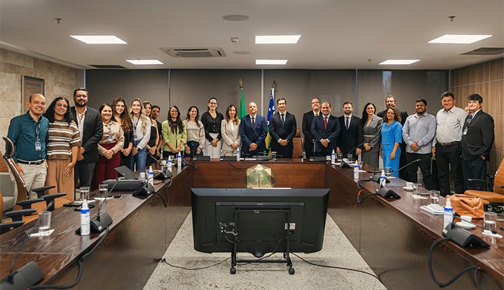 Imagem em formato horizontal de uma sala de reuniões institucional.  No centro, há uma grande mesa em formato retangular/ovalado, com cadeiras ao redor. Sobre a mesa estão microfones, copos de vidro com água, garrafas, álcool em gel e alguns papéis. Em primeiro plano, aparece um monitor grande voltado para o centro da mesa.  Ao fundo da sala, alinhadas lado a lado atrás da mesa principal, estão cerca de vinte pessoas posando para a foto. O grupo é composto por homens e mulheres adultos, vestindo trajes formais e sociais — ternos, gravatas, blazers, vestidos e camisas sociais. Alguns usam crachá de identificação. A maioria está sorrindo e olhando para a câmera.  No centro do grupo, dois homens de terno escuro e gravata ocupam posição de destaque, sugerindo possível papel de liderança. Ao fundo, atrás deles, há bandeiras oficiais posicionadas próximas à parede.  A sala tem iluminação embutida no teto, paredes em tons neutros e acabamento em madeira e pedra, transmitindo um ambiente formal e institucional.