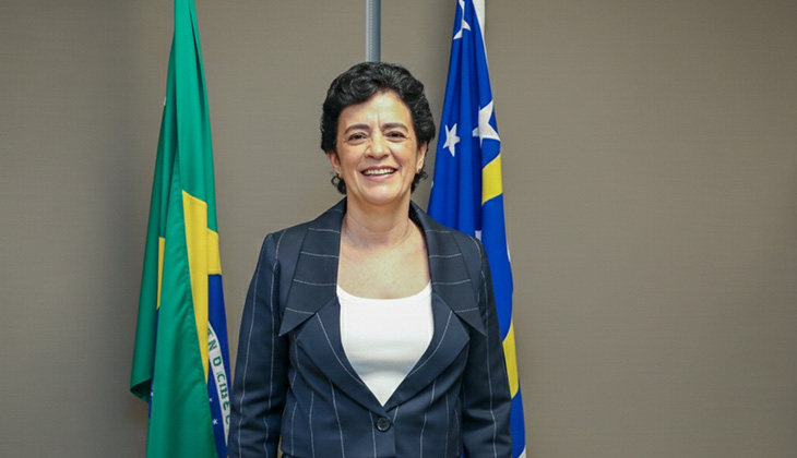A imagem mostra uma mulher em pé, posicionada de frente para a câmera, sorrindo. Ela tem cabelos curtos, escuros e levemente ondulados. Está vestindo um blazer escuro com listras finas claras e uma blusa branca por baixo. Usa brincos pequenos e discretos.  Ao fundo, há duas bandeiras: à esquerda, a bandeira do Brasil, com as cores verde, amarelo e azul; à direita, uma bandeira azul com estrelas e uma faixa amarela. O ambiente parece institucional ou oficial, com parede neutra e iluminação uniforme. A postura da mulher é ereta e confiante, transmitindo formalidade e cordialidade.
