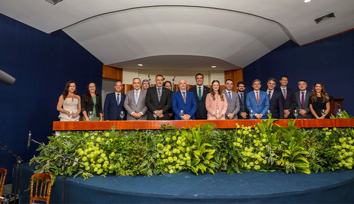 Fotografia horizontal registrada em ambiente interno durante uma solenidade. Em um palco elevado, cerca de quinze autoridades — homens e mulheres — estão alinhadas lado a lado atrás de uma mesa comprida de madeira. A maioria veste trajes formais: homens de terno e gravata e mulheres com vestidos ou ternos sociais. Sobre a mesa há copos de água e alguns microfones. À frente do palco, uma extensa decoração com plantas ornamentais verdes e flores claras cobre toda a parte frontal, formando um jardim decorativo. O fundo do palco é composto por paredes em tom azul-escuro e um teto curvo branco com iluminação embutida. No canto inferior esquerdo da imagem aparece parte de um microfone e de cadeiras, indicando área destinada ao público ou à imprensa. As autoridades posam para a foto de forma solene, voltadas para a câmera.