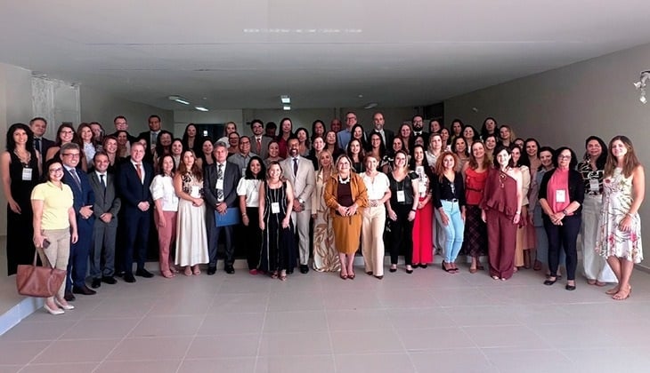 A imagem mostra um grande grupo de participantes reunidos para uma foto oficial em um espaço interno de um prédio institucional. Homens e mulheres, muitos usando crachá de identificação, estão organizados em várias fileiras e vestidos em trajes formais ou sociais. A fotografia registra os representantes de tribunais e instituições que participaram do encontro nacional sobre adoção e acolhimento promovido pelo Conselho Nacional de Justiça (CNJ).