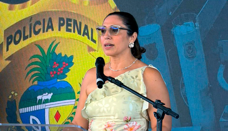 A imagem mostra a juíza Vaneska Baruki discursando em um púlpito transparente durante a cerimônia de inauguração da nova unidade prisional de Caldas Novas. Ela veste um vestido claro com estampa floral, óculos de sol e acessórios discretos. Ao fundo, há um grande painel com o símbolo da Polícia Penal.