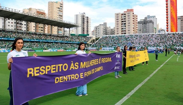 A imagem mostra pessoas posicionadas no gramado de um estádio de futebol segurando faixas de conscientização durante a final do Campeonato Goiano. Em destaque, uma faixa roxa com a mensagem “Respeito às mulheres é regra. Dentro e fora de campo”, reforçando a campanha de enfrentamento à violência contra a mulher. Ao fundo, as arquibancadas estão ocupadas por torcedores, evidenciando a realização da ação em meio ao evento esportivo.