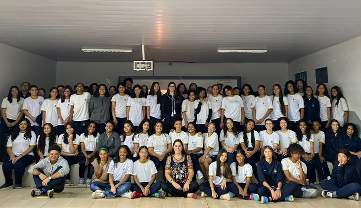 Imagem em formato horizontal mostra um grande grupo de pessoas reunidas em um ambiente interno, semelhante a uma sala escolar ou auditório. Ao todo, há dezenas de jovens, em sua maioria adolescentes, organizados em três fileiras: alguns sentados no chão à frente, outros sentados em cadeiras ou bancos no meio e um grupo maior em pé ao fundo.  A maioria veste camisetas brancas, possivelmente de uniforme escolar, combinadas com calças ou jeans. No centro da primeira fileira, destaca-se uma mulher adulta sentada no chão, usando blusa estampada e calça escura, sugerindo papel de liderança ou convidada especial. Outras duas mulheres adultas aparecem em pé ao centro da fileira de trás, também se destacando entre os estudantes.  Os jovens apresentam diversidade de tons de pele, cabelos e traços, e muitos sorriem ou mantêm expressões neutras, olhando diretamente para a câmera. O ambiente possui paredes claras, teto com iluminação fluorescente e, ao fundo, há um quadro escuro e um suporte de projetor fixado no teto.  A imagem transmite um clima coletivo, de integração e registro de um encontro ou atividade educacional