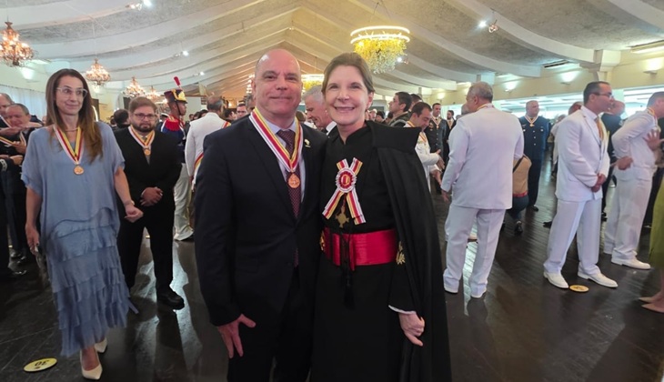 Em um salão amplo e bem iluminado, com teto decorado em arcos e lustres, várias pessoas participam de uma cerimônia formal. Em primeiro plano, ao centro, estão um homem e uma mulher posando lado a lado e sorrindo para a foto.  O homem é careca, de pele clara, e veste terno escuro com camisa branca e gravata. No peito, ele usa uma medalha pendurada por uma fita em tons de vermelho, amarelo e branco. A mulher, também de pele clara, tem cabelo castanho preso e veste uma toga preta com detalhes formais, além de uma faixa vermelha na cintura. No peito, ela também exibe uma insígnia com fita nas mesmas cores da medalha do homem.  Ao fundo, há diversas pessoas em trajes formais, incluindo alguns homens com uniformes brancos e outros participantes usando medalhas semelhantes, indicando tratar-se de uma solenidade oficial ou cerimônia de condecoração. O ambiente transmite um clima institucional e festivo.