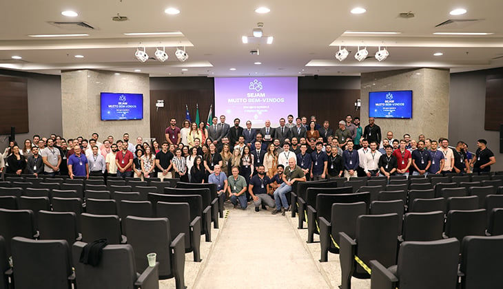 Imagem horizontal de um auditório moderno e bem iluminado. Em primeiro plano, aparecem várias fileiras de cadeiras pretas vazias, organizadas em formato de plateia e divididas por um corredor central.  Ao fundo, no palco, há um grande grupo de pessoas posando para uma foto oficial. São dezenas de participantes, homens e mulheres de diferentes idades e estilos, vestindo roupas formais e casuais, como camisas sociais, polos e blazers. A maioria está em pé, enquanto alguns estão agachados ou sentados na primeira fila para compor a foto.  No centro do palco, há uma tela grande com fundo roxo exibindo a mensagem “Sejam muito bem-vindos”, acompanhada de ícones gráficos. De cada lado da tela principal, há dois monitores menores com a mesma identidade visual. Atrás do grupo, também é possível ver bandeiras institucionais, indicando um evento oficial ou corporativo.  O ambiente transmite a ideia de um encontro institucional, treinamento ou evento corporativo, com organização formal e clima de integração entre os participantes.