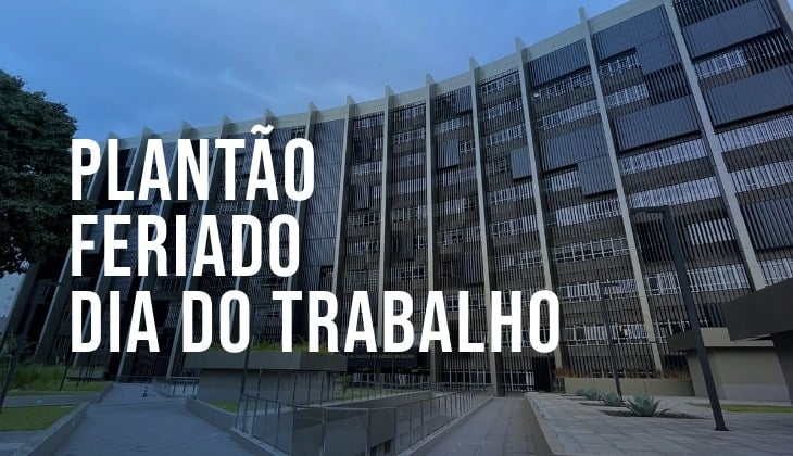 Imagem em formato horizontal mostra a fachada de um prédio institucional de grande porte, com vários andares e estrutura moderna, composta por brises verticais e janelas alinhadas. A construção ocupa a maior parte do enquadramento e é vista a partir de um ângulo levemente baixo, destacando sua altura.  À esquerda, há uma área com gramado e uma árvore. À frente do edifício, vê-se um caminho pavimentado que leva à entrada principal, com rampas e corrimãos metálicos. A iluminação sugere período diurno, com céu parcialmente nublado.  Sobre a imagem, em destaque, há um texto em letras grandes, brancas e em caixa alta, disposto em três linhas: “PLANTÃO”, “FERIADO” e “DIA DO TRABALHO”. O texto ocupa a parte esquerda e central da imagem, contrastando com o fundo escuro do prédio.