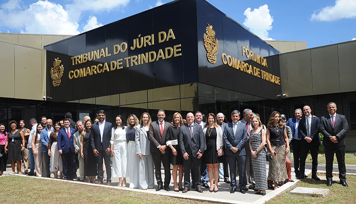 Imagem colorida, em formato horizontal, registrada em área externa durante o dia. Em primeiro plano, um grupo numeroso de pessoas — homens e mulheres — está posicionado lado a lado, formando uma fileira. Todos estão trajando roupas formais, como ternos, vestidos e conjuntos sociais, em tons variados, predominando preto, cinza e branco. A maioria sorri e olha em direção à câmera, transmitindo um clima institucional e de celebração.  Ao fundo, destaca-se a fachada moderna de um prédio público, com revestimento escuro e letras em destaque na cor dourada. É possível ler as inscrições “Tribunal do Júri da Comarca de Trindade” e “Fórum da Comarca de Trindade”, além de brasões institucionais. A construção possui linhas retas e acabamento contemporâneo.  O céu está azul, com poucas nuvens, e a iluminação natural é forte, indicando um dia ensolarado. A área ao redor apresenta gramado bem cuidado, reforçando o ambiente organizado e institucional.