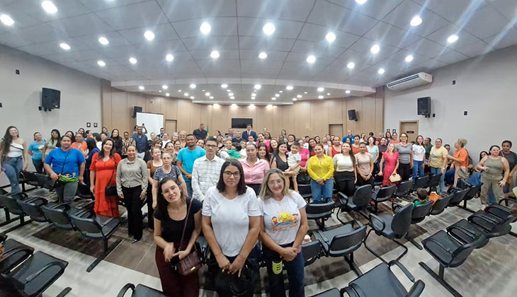 A imagem mostra um auditório bem iluminado, com dezenas de pessoas reunidas em pé entre as fileiras de cadeiras. O grupo é diverso, composto majoritariamente por adultos, como em uma foto coletiva ao final de um evento. Ao fundo, há paredes revestidas em madeira, equipamentos de som e um ar-condicionado.