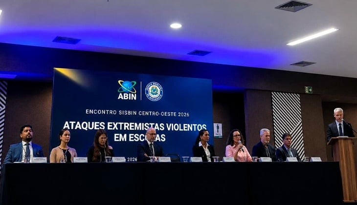 A imagem de evento institucional realizado em auditório, com uma mesa composta por várias autoridades sentadas lado a lado. Ao fundo, há um painel azul com os logotipos da Abin e do Sisbin e o título do encontro: “Ataques extremistas violentos em escolas”, indicando o tema central da discussão. À direita, um homem está em pé, falando ao púlpito, enquanto os demais participantes acompanham. A iluminação é moderna, com luzes no teto, e o ambiente transmite formalidade e caráter oficial. 