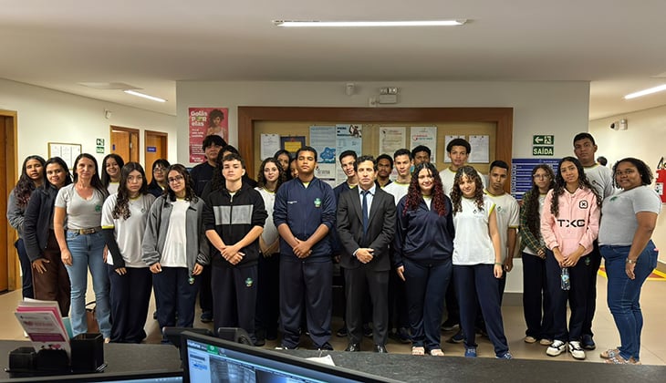 A imagem mostra um grupo de adolescentes, em sua maioria uniformizados, acompanhados por alguns adultos, posando para uma foto em um ambiente institucional interno. No centro, há um homem de terno, em posição de destaque. O grupo está organizado em filas, com expressão neutra ou leve sorriso, voltado para a câmera. Ao fundo, há um mural com avisos e cartazes, além de sinalizações de saída de emergência nas laterais. Em primeiro plano, aparecem parcialmente monitores de computador, indicando que a foto foi feita em um espaço institucional. 