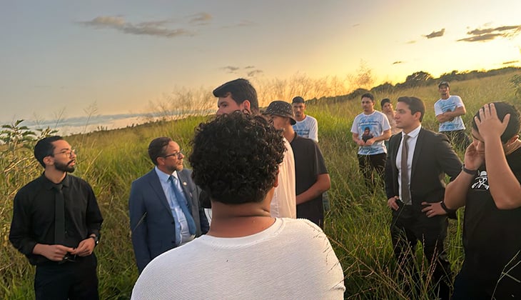 A imagem mostra um grupo de pessoas reunidas em uma área de vegetação alta, ao ar livre, durante o fim da tarde, com o céu iluminado por uma luz dourada. Algumas pessoas estão vestidas formalmente, como ternos e camisa social, enquanto outras usam roupas casuais.