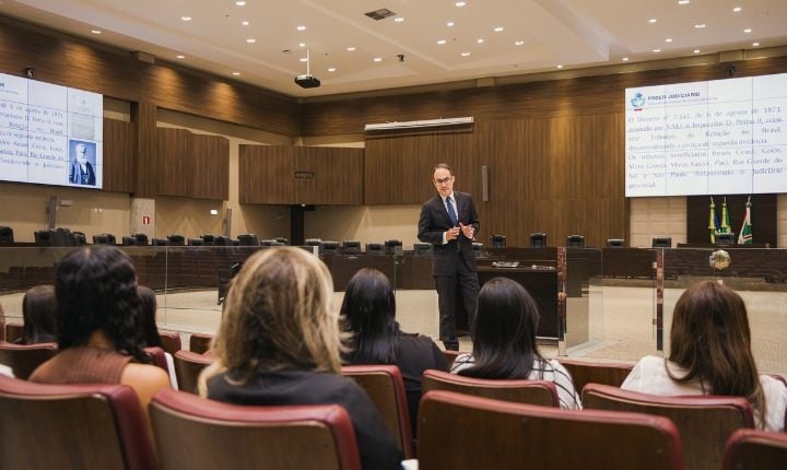  A imagem mostra um ambiente formal, como um auditório de um tribunal. Um homem de terno está em pé, à frente, fazendo uma apresentação. Ele parece estar explicando algo para um grupo de pessoas sentadas em cadeiras, que o observam atentamente. O espaço tem decoração moderna, com paredes de madeira e iluminação embutida no teto. Ao fundo, há uma mesa grande e bandeiras, reforçando a ideia de um ambiente institucional ou jurídico. Também é possível ver telas projetando conteúdo, indicando que a apresentação utiliza recursos visuais.