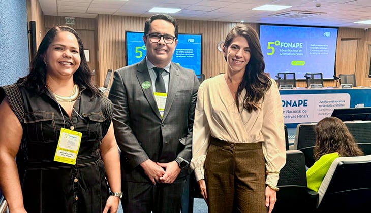 A imagem mostra três pessoas posando para foto em um auditório no qual se encontram outras pessoas. Ao fundo, aparecem uma mesa que sugere a ideia de uma conferência, telas azuis e um painel com o nome “5º Fonape”
