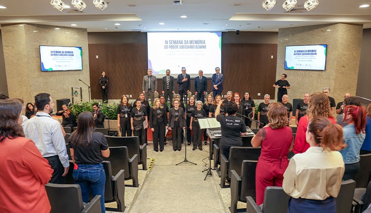 Descrição da imagem: Fotografia de um auditório durante uma apresentação musical. À frente, um coral formado por várias pessoas vestidas de preto canta em pé, posicionado diante do palco. No centro, uma regente aparece de costas, conduzindo o grupo. Há microfones, teclado e outros equipamentos musicais no espaço.  Ao fundo, no palco, algumas autoridades estão em pé, usando trajes formais. Nas laterais do auditório, duas telas exibem a identidade visual do evento “IV Semana da Memória do Poder Judiciário Goiano”. Na plateia, várias pessoas acompanham a apresentação, algumas em pé entre as fileiras de cadeiras. O ambiente é iluminado e tem paredes em tons claros e amadeirados.