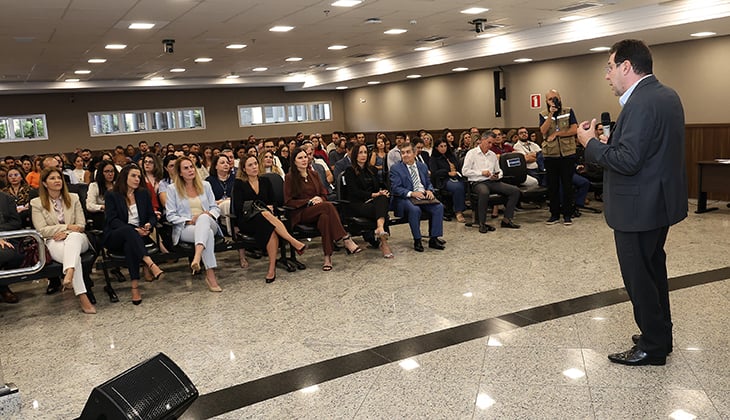 Fotografia mostra um auditório amplo e iluminado durante uma palestra ou evento institucional. Em primeiro plano, à direita, um homem de terno escuro está em pé, de costas para a câmera, falando ao público com um microfone na mão. À frente dele, dezenas de pessoas estão sentadas em cadeiras organizadas em fileiras, acompanhando atentamente a apresentação. A plateia é composta por homens e mulheres vestidos com trajes sociais e executivos. O ambiente possui piso claro e brilhante, teto com iluminação embutida e paredes em tons neutros. Ao fundo, há pequenas janelas horizontais e uma profissional registrando o momento com câmera fotográfica.