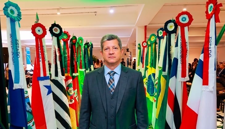  Um homem de terno cinza e gravata está posicionado ao centro da imagem, olhando diretamente para a câmera com expressão neutra. Ao seu redor, há diversas bandeiras de estados brasileiros, em fileiras, adornadas com rosetas coloridas no topo. O ambiente é a abertura do 6º Enam, no Tribunal de Justiça do Pará.