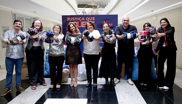 Em um ambiente interno amplo e iluminado, nove pessoas estão posicionadas lado a lado, sorrindo e segurando cobertores enrolados em direção à câmera. Ao fundo, há um painel azul com a frase “Justiça que Aquece” em destaque, além de ilustrações relacionadas ao inverno. Atrás do grupo, aparecem várias sacolas e pacotes empilhados, sugerindo uma campanha de arrecadação de agasalhos e cobertores. O espaço possui piso em tons de preto e branco e arquitetura moderna, com corredores e paredes claras ao redor.