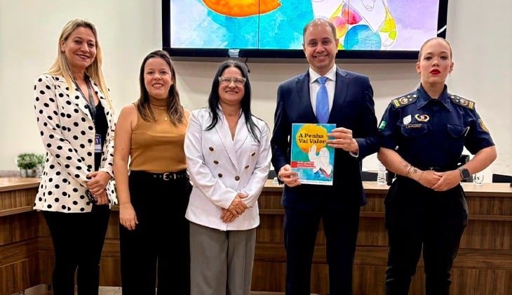 A imagem mostra cinco pessoas em pé, lado a lado, em um ambiente interno. Ao centro, um homem de terno segura um livro colorido onde se lê o título "A Penha Vai Valer". À esquerda dele estão três mulheres com roupas sociais e, à direita, uma mulher vestindo uniforme oficial. 