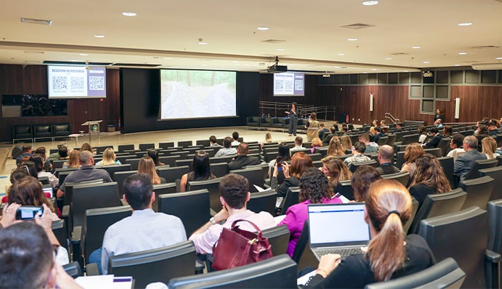 Fotografia colorida de um auditório de grande porte durante uma palestra ou evento institucional. A imagem foi registrada da parte posterior da plateia, mostrando dezenas de participantes sentados em poltronas estofadas, voltados para o palco. Algumas pessoas utilizam notebooks, celulares ou fazem anotações, enquanto outras acompanham a apresentação.  À frente, um palestrante está em pé no palco, próximo a uma grande tela de projeção que exibe a imagem de uma estrada cercada por vegetação. Em ambos os lados do palco há telões menores com um QR Code e informações da apresentação. O palco também conta com uma mesa, cadeiras e um púlpito.  O auditório possui paredes revestidas em madeira escura, iluminação embutida no teto e corredores amplos entre as fileiras de assentos. O ambiente transmite o contexto de um seminário, oficina ou capacitação, com os participantes atentos à exposição.
