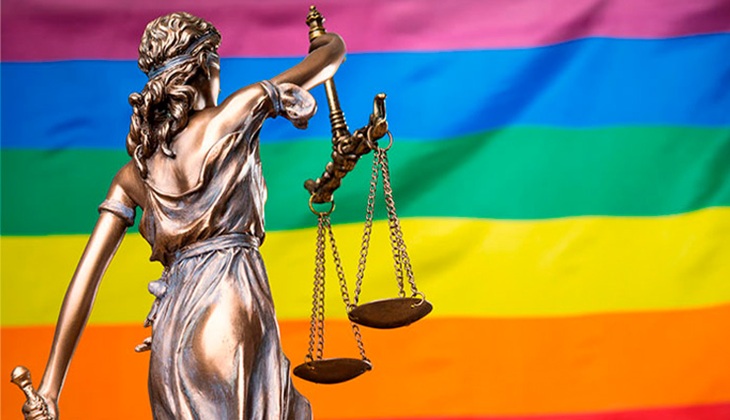 Imagem mostra, em primeiro plano, uma escultura clássica da deusa da Justiça (Themis), em tom metálico bronzeado. A figura está de costas e levemente de lado, com o braço direito erguido segurando uma balança de dois pratos, símbolo de equilíbrio e न्याय. No braço esquerdo, parcialmente visível, ela segura uma espada apontada para baixo.  Ao fundo, ocupando toda a imagem, há uma bandeira com listras horizontais nas cores do arco-íris (vermelho, laranja, amarelo, verde, azul e roxo), associada à diversidade e à comunidade LGBTQIA+. O contraste entre a estátua clássica e a bandeira colorida sugere a relação entre justiça e direitos das pessoas LGBTQIA+